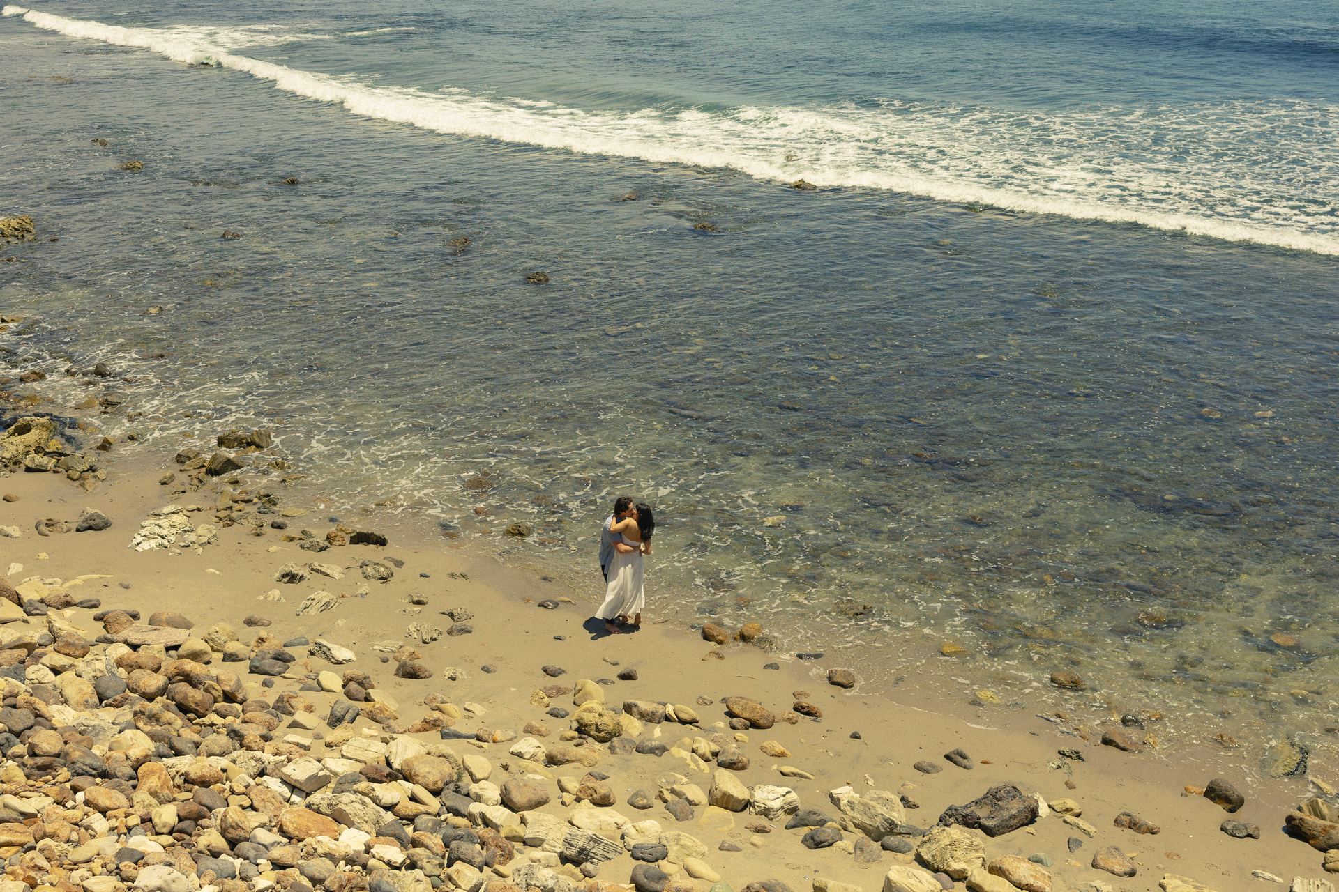 Grace and Zach embracing on a rocky beach at the water's edge